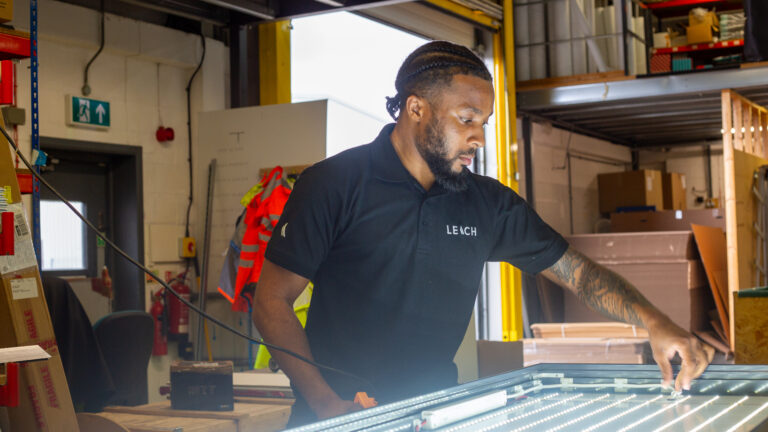 Leach employee wearing branded t-shirt works on producing a lightbox, in the background there is a factory.