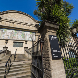 Signage mounted by the entrance to the museum, it is black with white writing. In the background there are stairs, lamps and museum's building with fresco painting